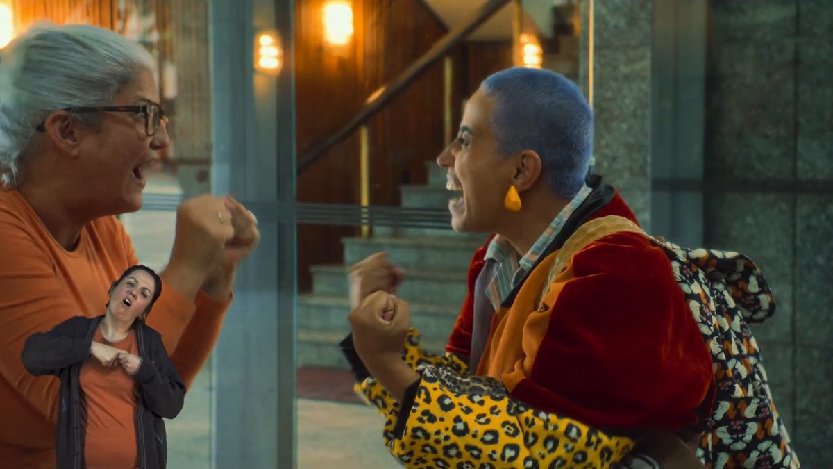Cena de “Nem Toda História de Amor Acaba em Morte”: duas personagens conversam animadas enquanto uma intérprete de Libras, dentro da cena, sinaliza a fala