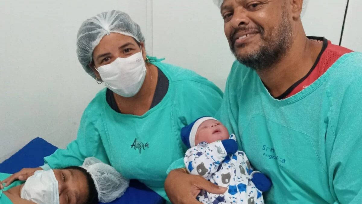 A mãe Débora está deitada na maca; a intérprete Thayane, em pé, a abraça, enquanto o pai Nélson acompanha ao lado