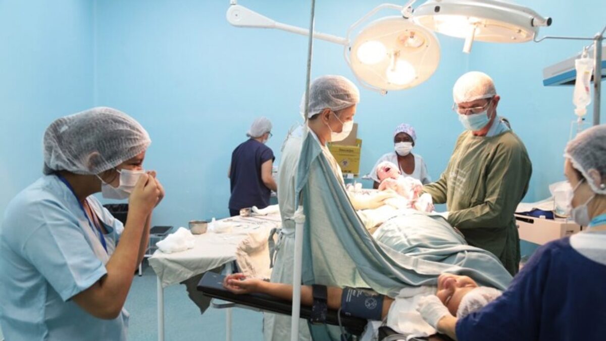 Sala de cirurgia; em primeiro plano, a intérprete faz sinais para a mãe, enquanto ao fundo a equipe médica segura o bebê