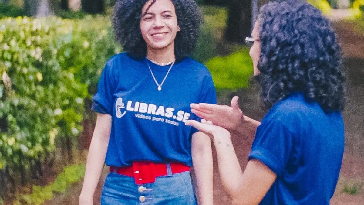 Duas pessoas com camisetas da LIBRAS.SE conversando em Libras ao ar livre