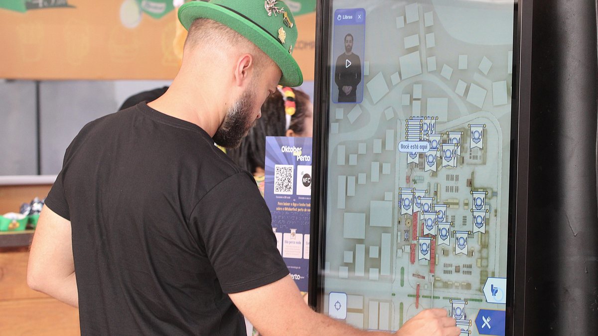 Visitante de chapéu verde usa o totem do aplicativo da Oktoberfest, com o mapa da festa e a janela de Libras na tela