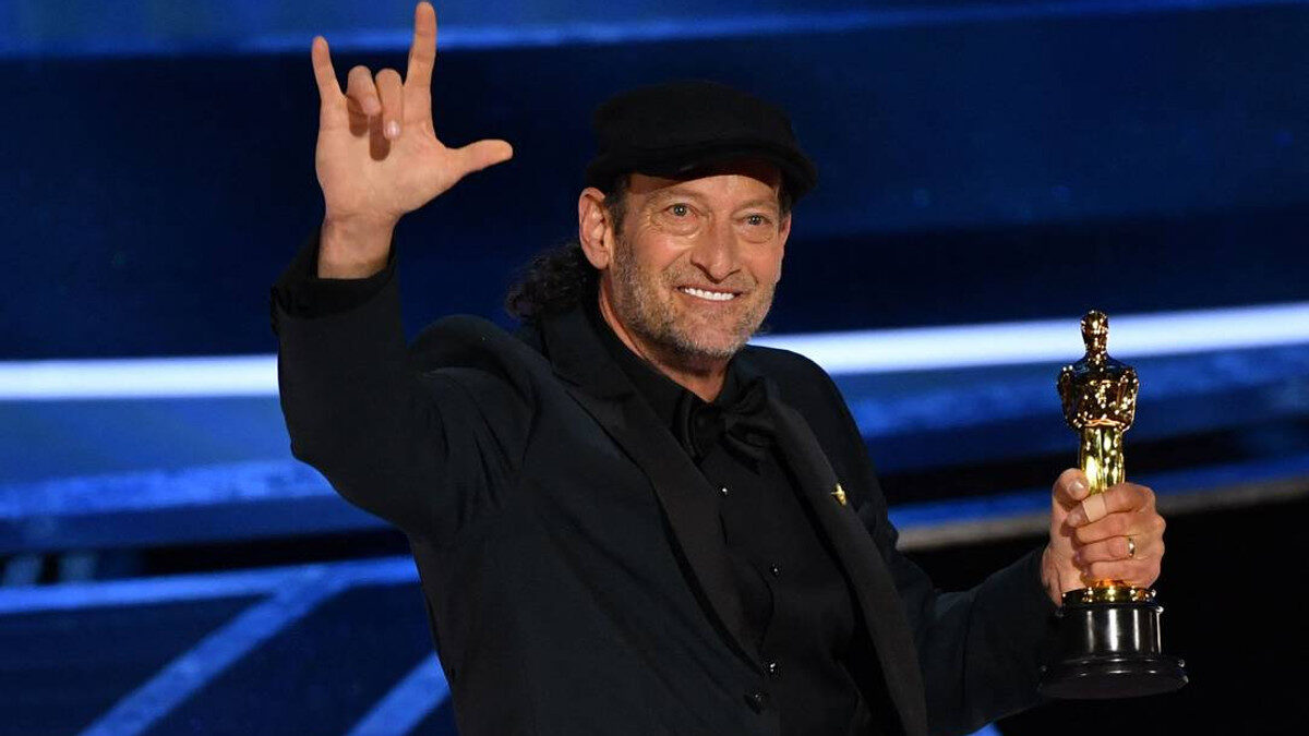 Troy Kotsur, de terno e chapéu, sorri e faz o sinal de “eu te amo” enquanto segura sua estatueta do Oscar
